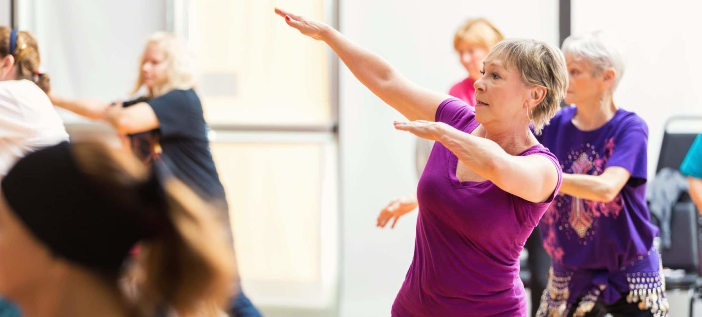 Diverse group of senior women take dance lessons in exercise studio at senior center. The women have their arms up as they learn the dance. Large bright windows are in the background.