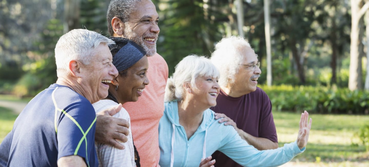 A group of five multi-ethnic seniors standing together in a park wearing casual clothing, looking to the side, smiling. The Caucasian woman wearing light blue is waving at someone.