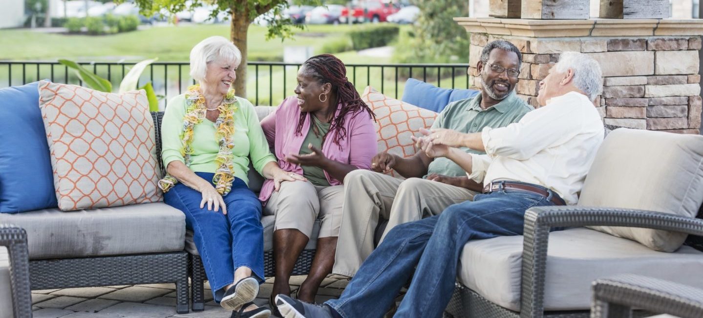 A multi-ethnic group of mature and senior friends hanging out together, sitting outdoors on a patio conversing. The African-American couple are in their 50s. The Caucasian couple are in their 70s. The women are sitting next to each other, talking, and the men are chatting with each other.